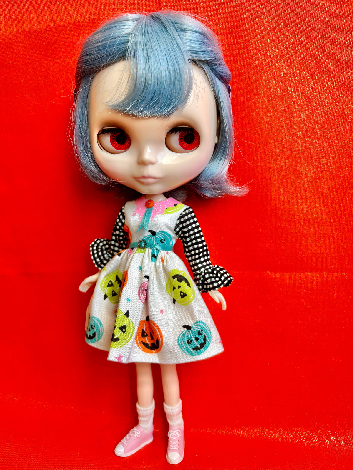 A stylized doll with large red eyes, short blue hair, and a white dress decorated with colorful jack-o'-lantern faces, standing against a bright red background.