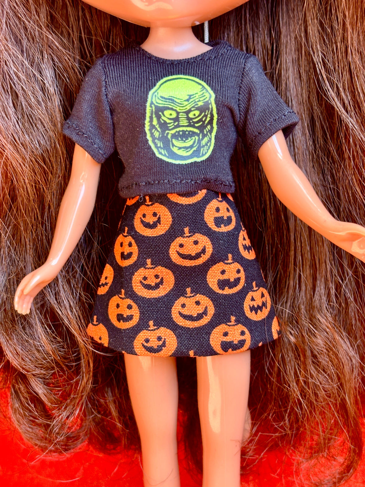 A doll with long brown hair wearing a dark gray short-sleeve knit top with a green zombie face graphic and a black skirt with an orange pumpkin print, set against a background of brown hair and a red surface.