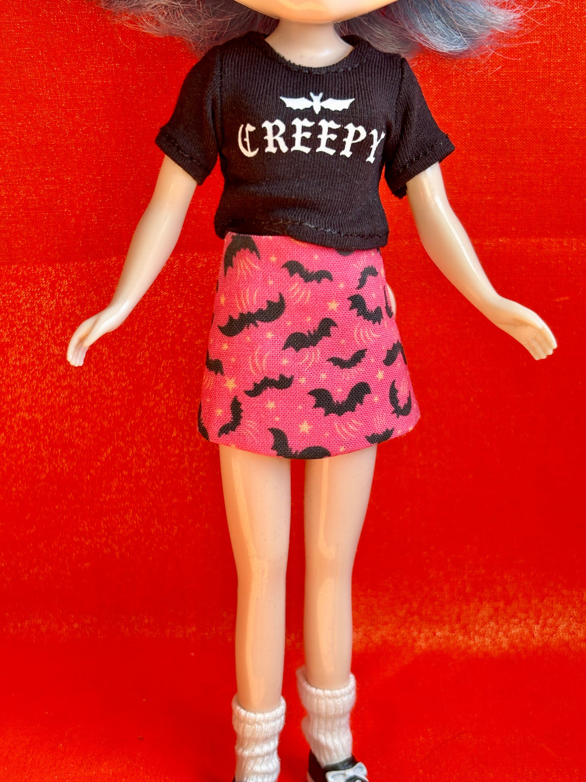 A doll or mannequin wearing a black short-sleeve t-shirt with 'CREEPY' and bat graphics, paired with a pink skirt patterned with black bats and small stars, standing against a bright red background.