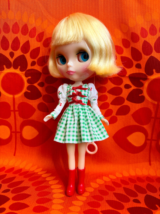 Doll with blonde hair, blue eyes, and a green checkered dress against an orange patterned background.