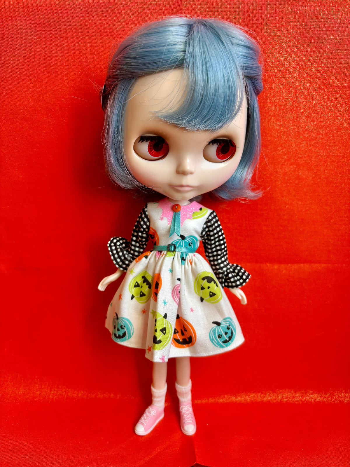 A stylized doll with large red eyes, short blue hair, and a Halloween-themed dress featuring pumpkins, shown against a bright red background.