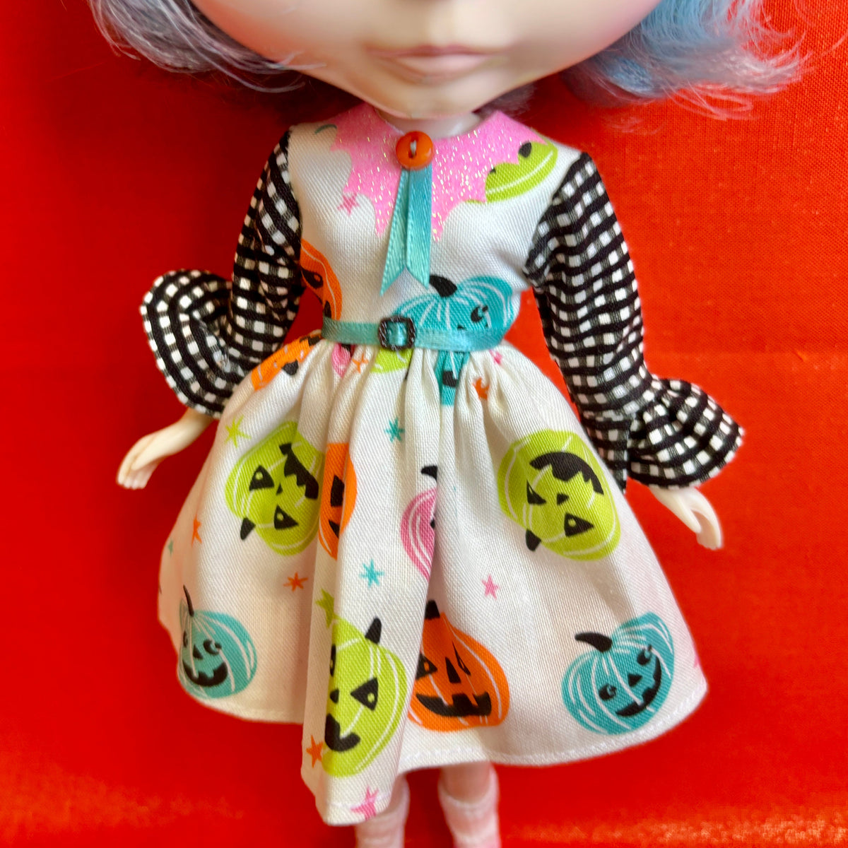 A close-up of a doll wearing a Halloween-themed dress with a cream base, black-and-white checkered sleeves, and colorful jack-o'-lantern prints, posed against a bright red background.