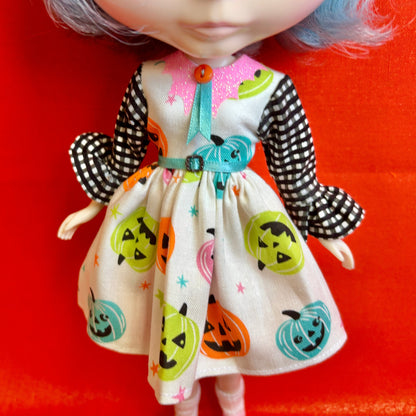 A close-up of a doll wearing a Halloween-themed dress with a cream base, black-and-white checkered sleeves, and colorful jack-o'-lantern prints, posed against a bright red background.