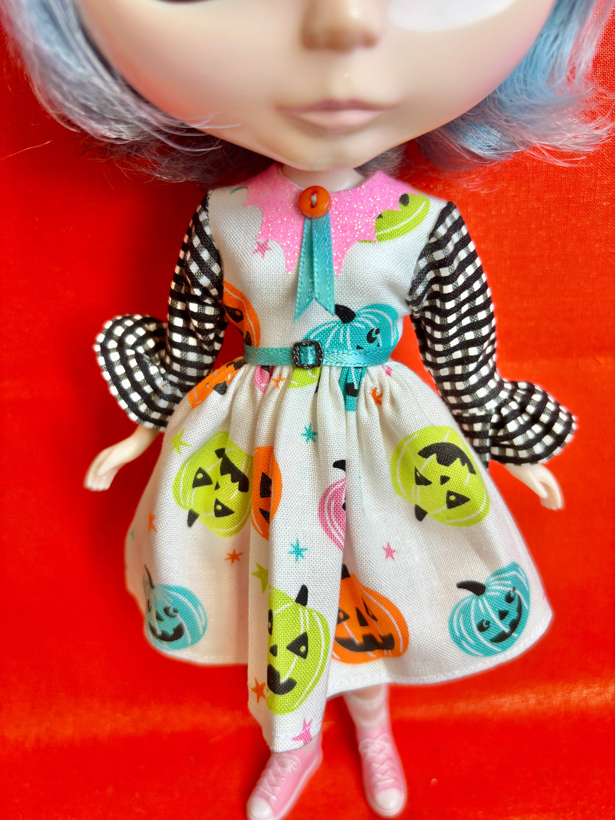 A doll wearing a Halloween-themed dress with a white base, black-and-white checkered sleeves, a pink collar, and colorful jack-o'-lantern prints, posed against a bright red background.
