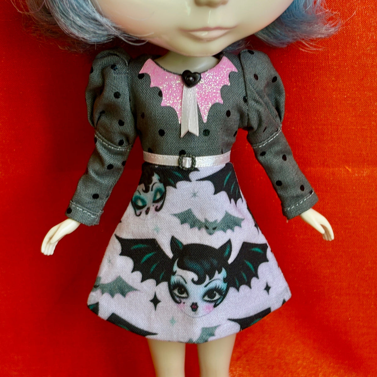 A doll wearing a gray long-sleeve dress with black polka dots, a pink glitter bat collar, and a printed skirt featuring cartoon bats on a white cloud pattern, posed against a bright red-orange background.