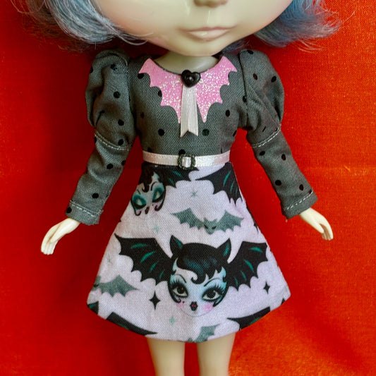 A doll wearing a gray long-sleeve dress with black polka dots, a pink glitter bat collar, and a printed skirt featuring cartoon bats on a white cloud pattern, posed against a bright red-orange background.