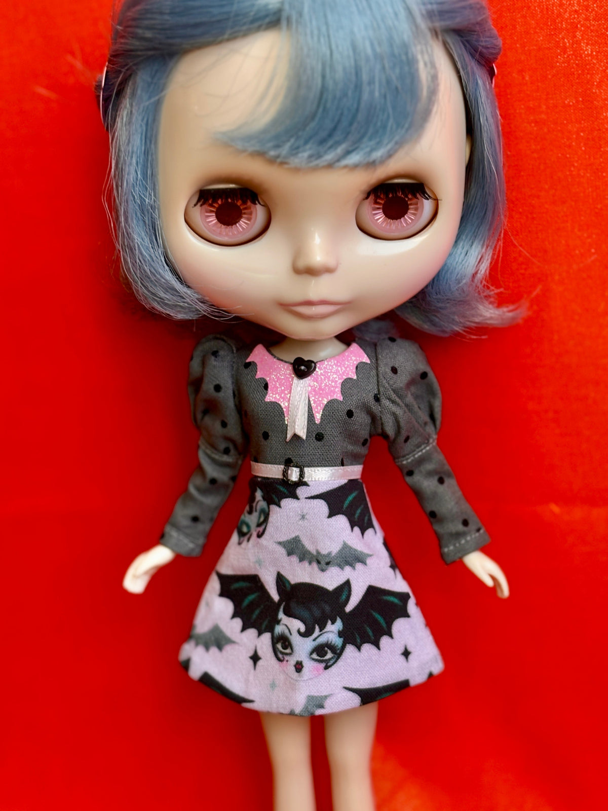 A Blythe-style doll with large pink eyes and blue hair, wearing a dark gray long-sleeve dress with a pink bat collar and a light pink skirt printed with black bats and a vampire face, posed against a bright red background.