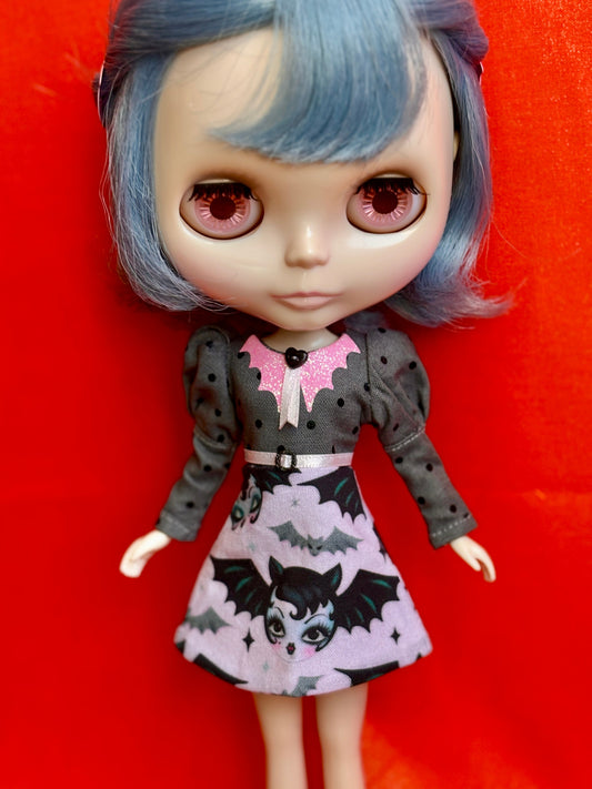 A Blythe-style doll with large pink eyes and blue hair, wearing a dark gray long-sleeve dress with a pink bat collar and a light pink skirt printed with black bats and a vampire face, posed against a bright red background.