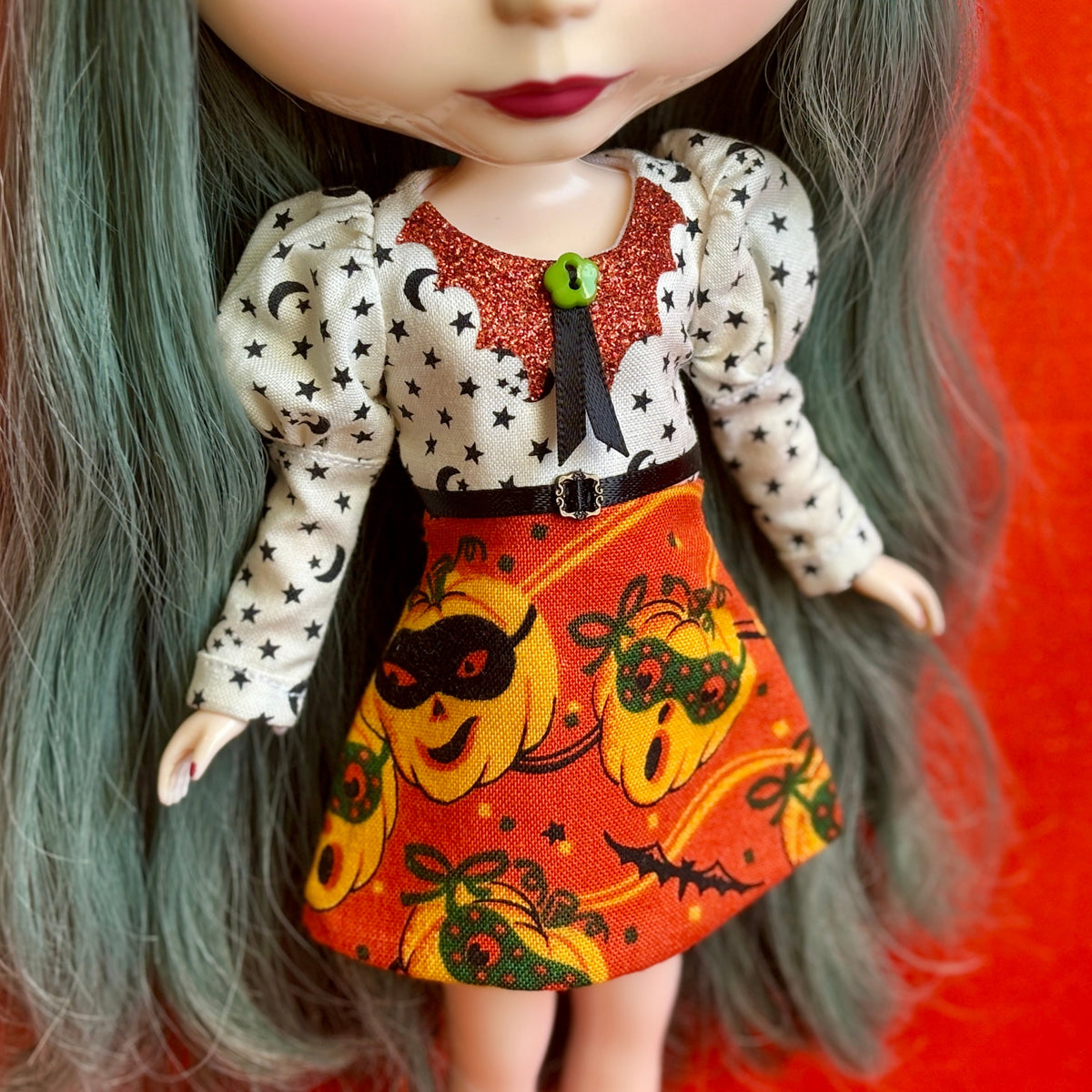 A close-up of a doll wearing a Halloween-themed dress with a white long-sleeve top patterned with black stars and crescent moons, a glittery red collar, a black ribbon tie, and an orange skirt with a pumpkin print.