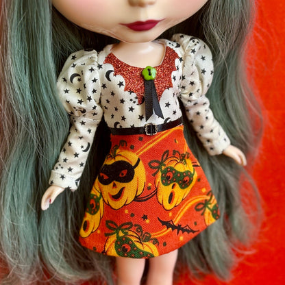 A close-up of a doll wearing a Halloween-themed dress with a white long-sleeve top patterned with black stars and crescent moons, a glittery red collar, a black ribbon tie, and an orange skirt with a pumpkin print.