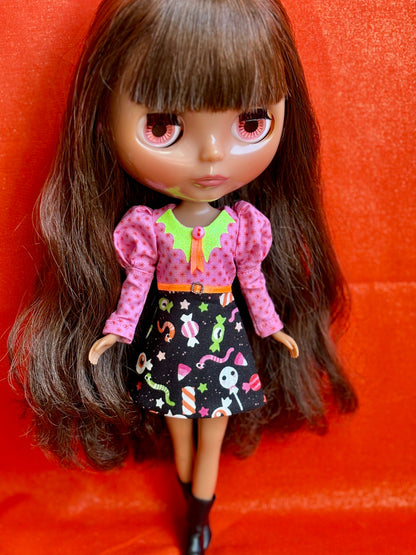 A doll with long brown hair and large pink eyes, wearing a pink long-sleeve top with a green collar and a black skirt with colorful candy prints, posed against a bright orange-red textured background.