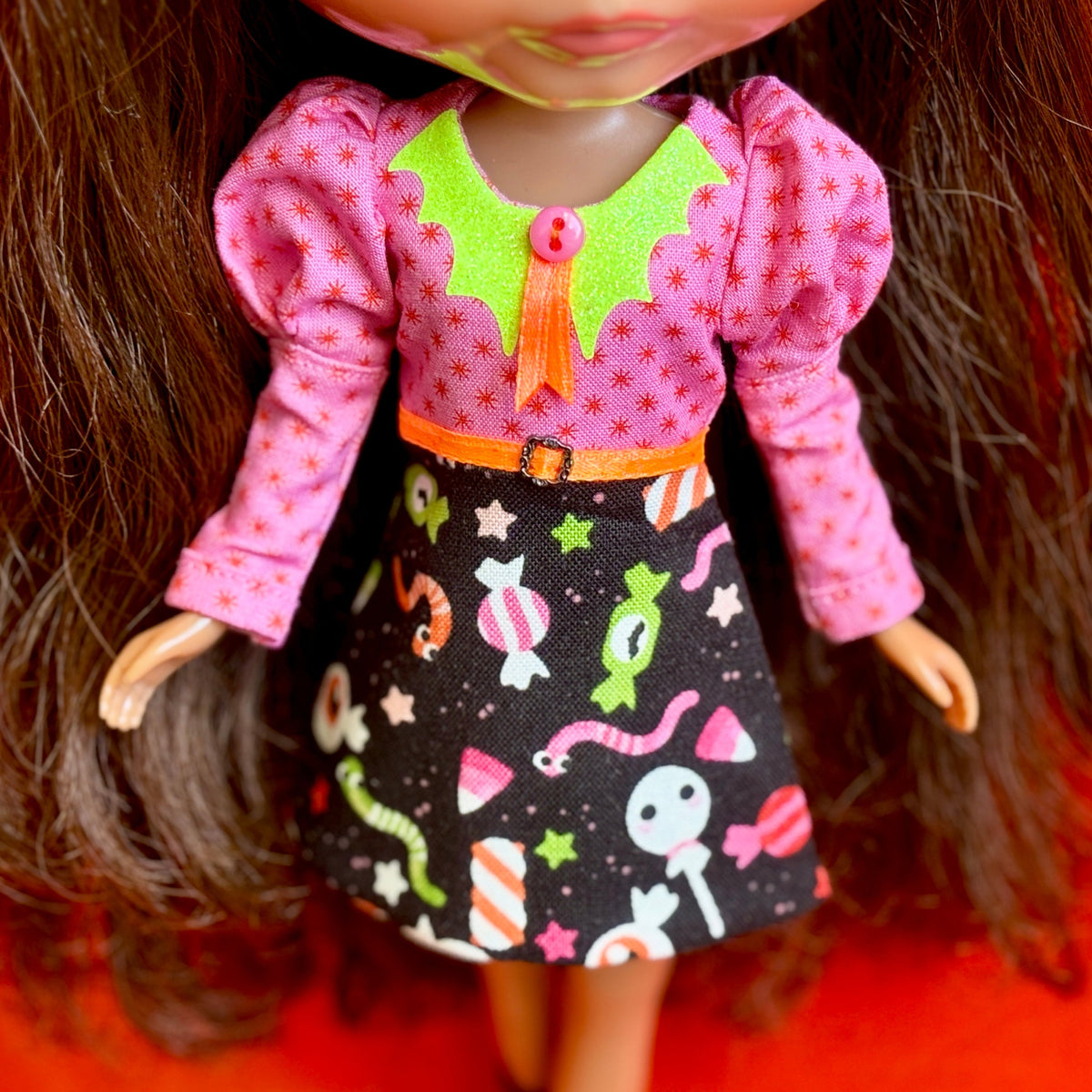 A close-up of a doll wearing a pink long-sleeve top with a lime green collar, an orange button and bow, and a black skirt printed with colorful candy and worm motifs.