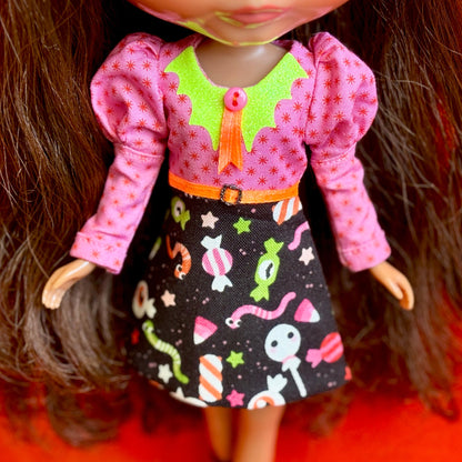 A close-up of a doll wearing a pink long-sleeve top with a lime green collar, an orange button and bow, and a black skirt printed with colorful candy and worm motifs.