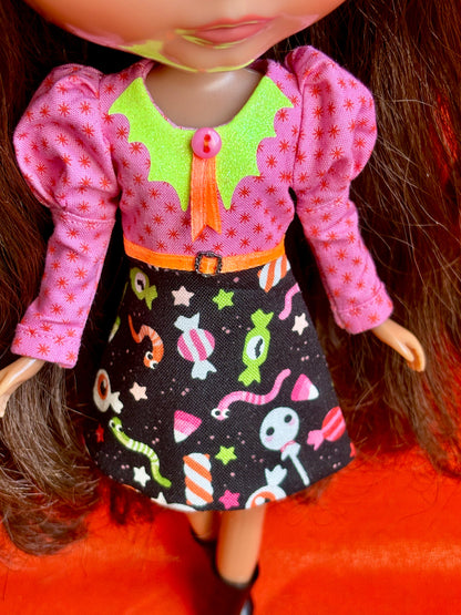 A close-up of a doll wearing a colorful costume dress with a pink patterned top, lime green collar, orange bow, and a black skirt with candy and worm prints, set against a bright orange background.
