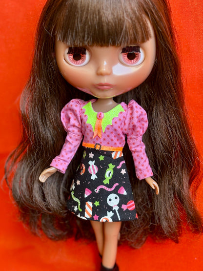 A close-up of a doll with long dark brown hair and large pink eyes, wearing a pink long-sleeve top with a green collar and a black skirt with colorful candy and ghost patterns, posed against a bright orange-red background.