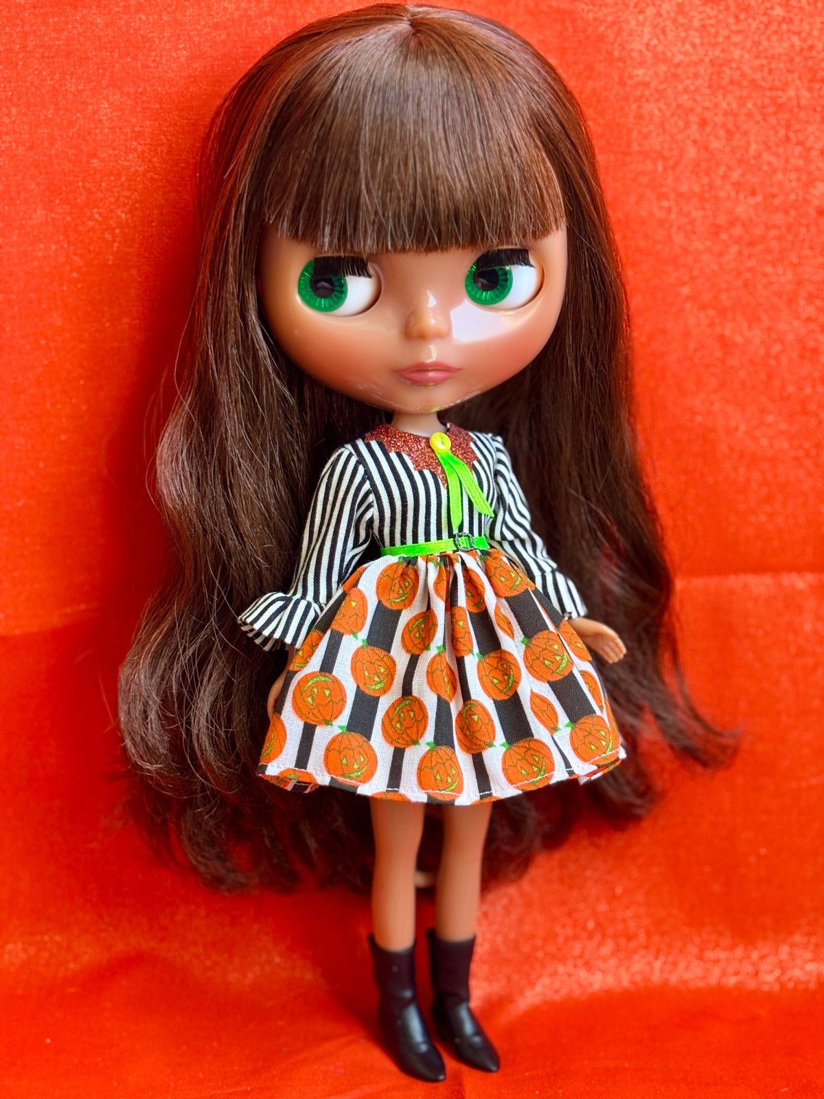 A Blythe-style doll with long brown hair and green eyes, wearing a black-and-white striped top and an orange, black, and white patterned skirt, standing against a bright orange-red fabric background.