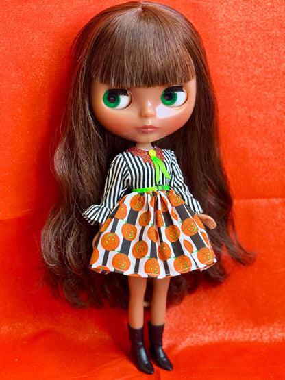 A Blythe-style doll with long brown hair and green eyes, wearing a black-and-white striped top and an orange, black, and white patterned skirt, standing against a bright orange-red fabric background.