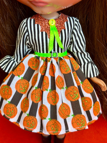 A doll wearing a Halloween-themed dress with a black and white striped long-sleeve top, a glittery orange collar, a bright green bow, and a white skirt printed with orange jack-o'-lanterns and dark brown vertical stripes.