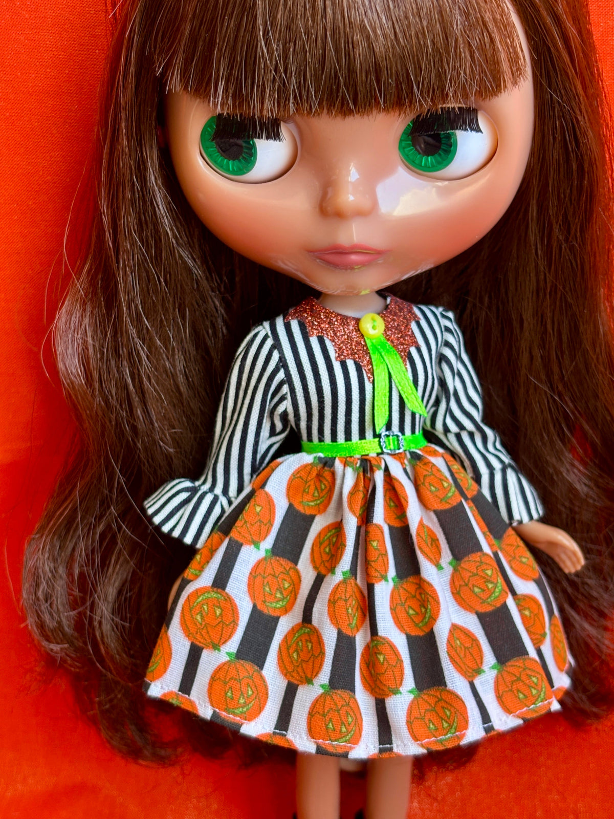 A close-up of a doll with large green eyes and long brown hair, wearing a Halloween-themed dress with a black and white striped long-sleeve top and a white skirt patterned with orange pumpkins, set against a bright orange background.