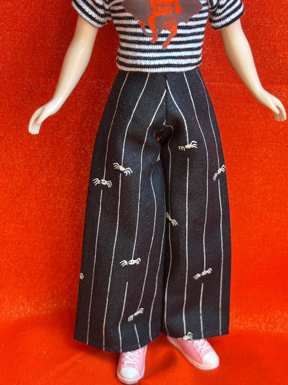 A doll or mannequin wearing a black-and-white striped short-sleeve top with an orange spider graphic, black pinstriped wide-leg pants with small white spider motifs, and pink shoes, posed against a bright orange-red textured backdrop.