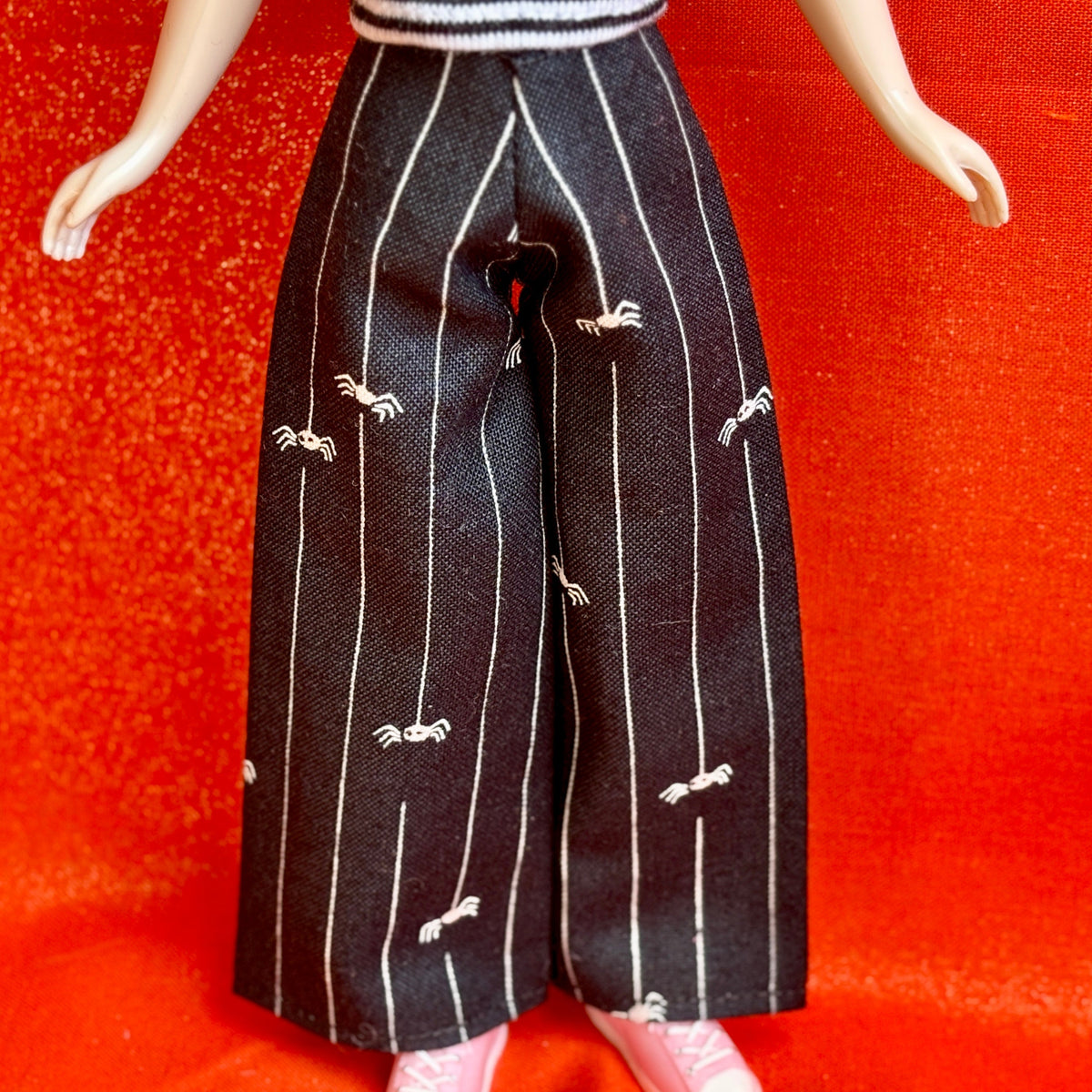 Black wide-leg pants with white vertical pinstripes and small white spider motifs, shown on a doll against a bright red textured background.