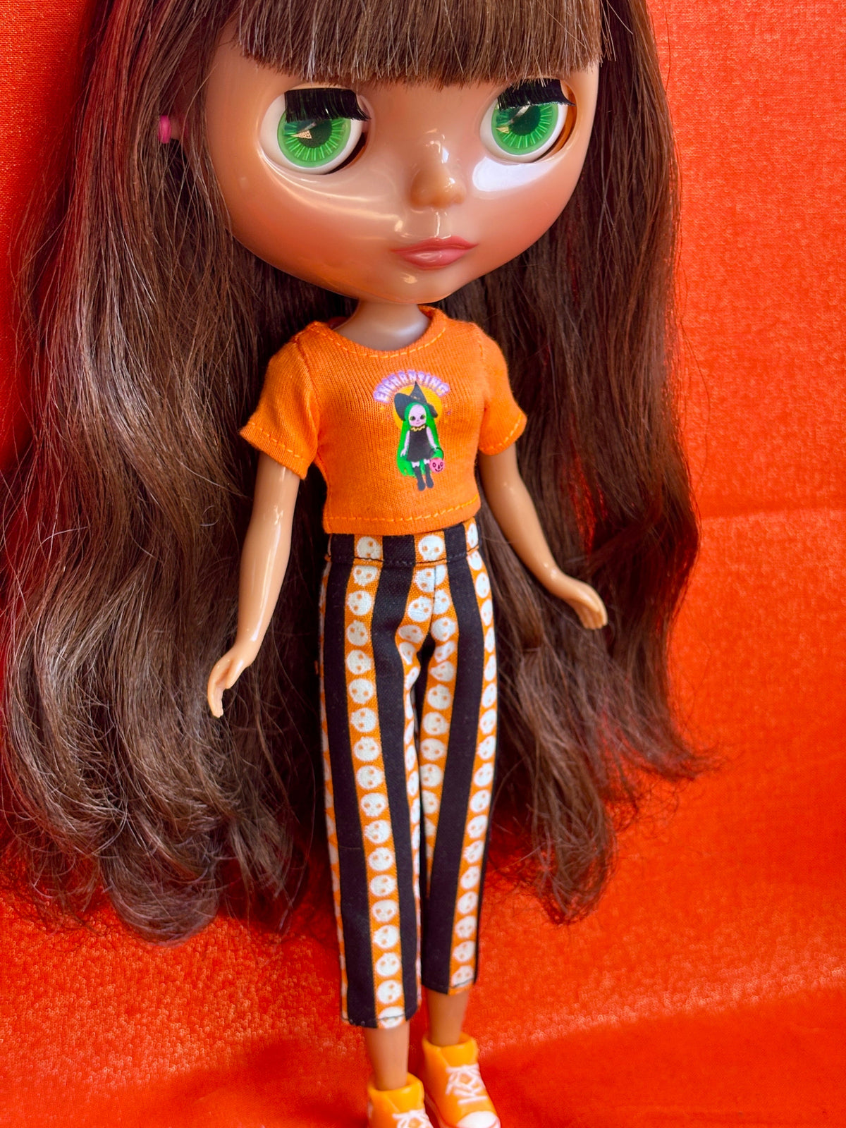 A doll with long brown hair and large green eyes, wearing an orange graphic T-shirt and black-and-white patterned pants, posed against a bright orange background.