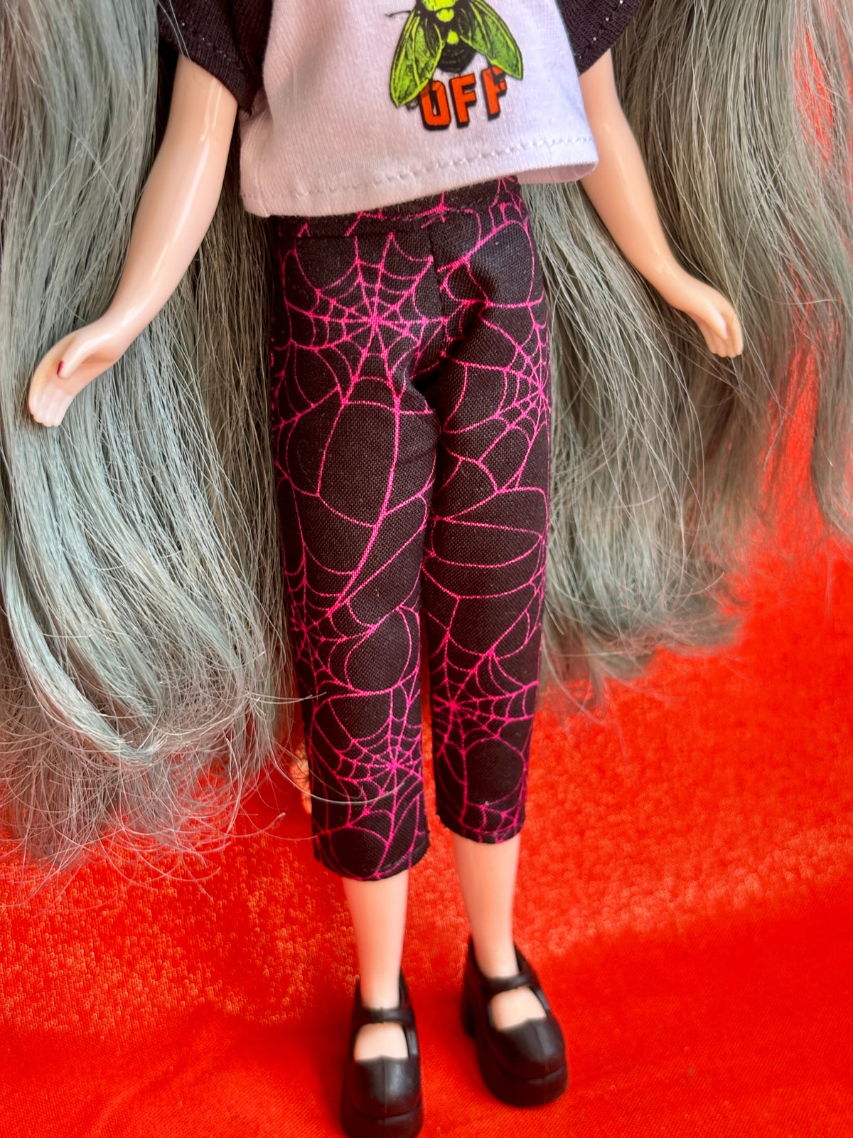 A doll or fashion figure shown from the waist down, wearing black cropped pants with a bright pink spiderweb pattern, a light gray graphic top, and black shoes, posed against a vivid red background.
