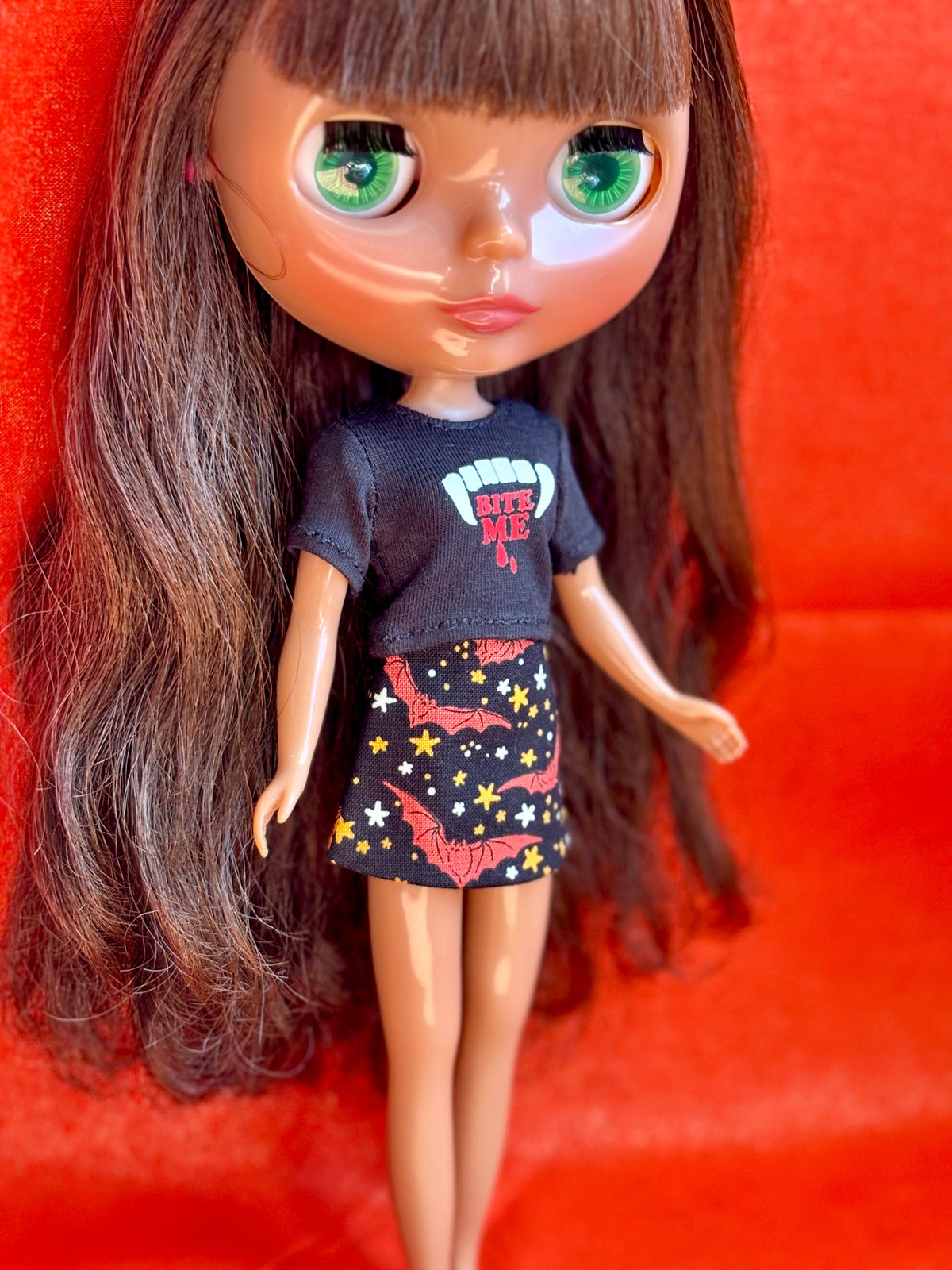 A close-up of a doll with long brown hair and large green eyes, wearing a black graphic T-shirt and a black skirt with red and yellow star-and-bat print, posed against a bright red background.
