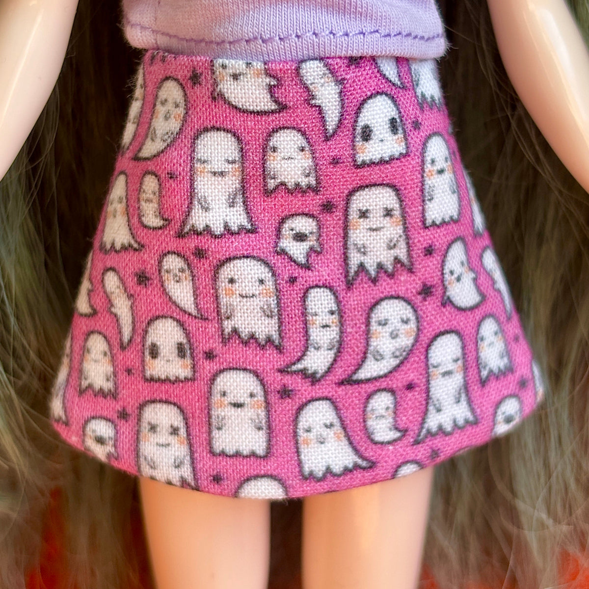 A close-up of a doll wearing a pink skirt with a repeating pattern of cute white cartoon ghosts and small black stars.