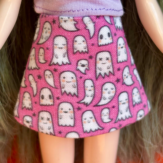 A close-up of a doll wearing a pink skirt with a repeating pattern of cute white cartoon ghosts and small black stars.