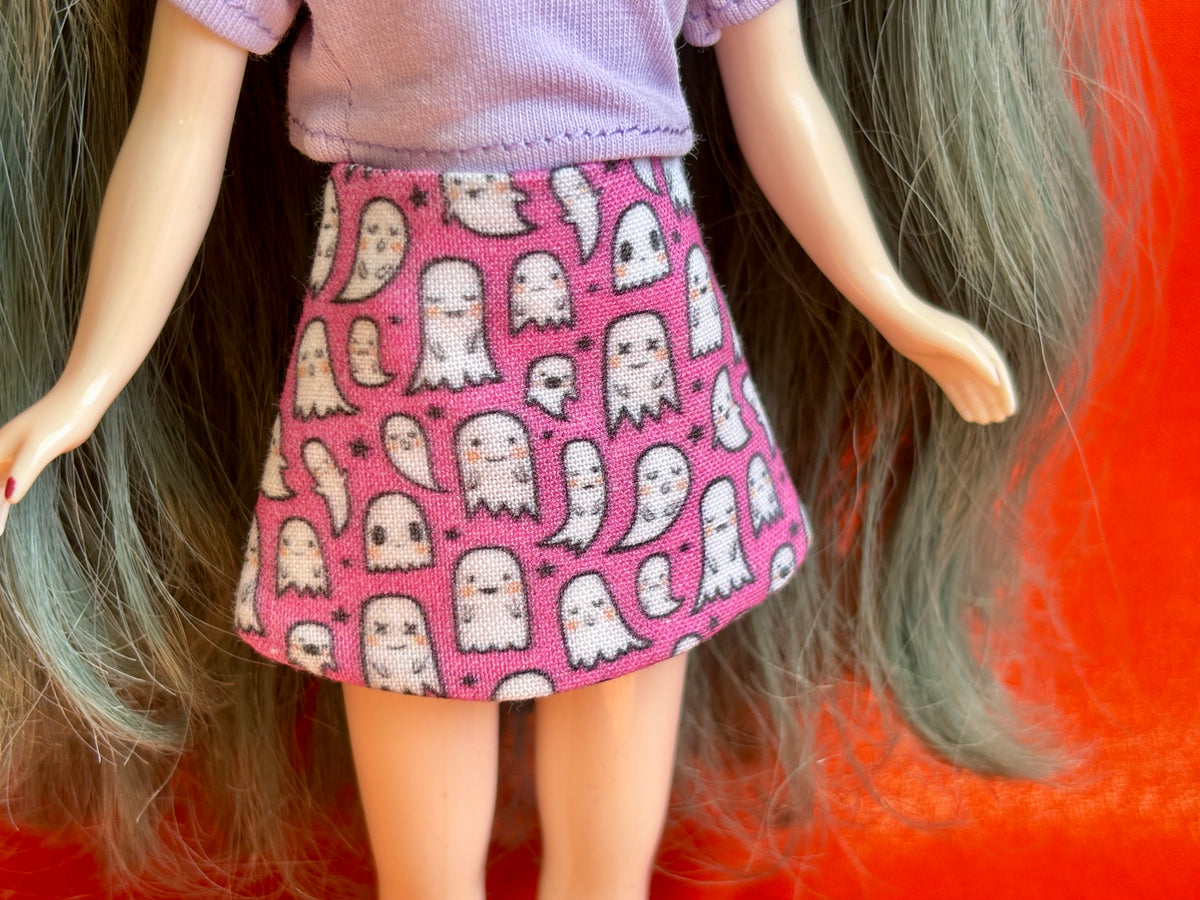 A close-up of a doll wearing a pink skirt with a repeating white ghost pattern, paired with a lavender top, set against a bright orange background.