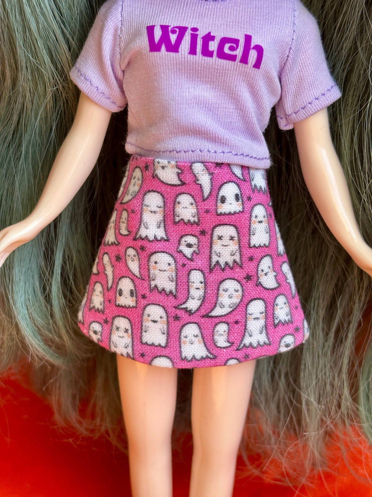 A doll or toy figure shown from the back, wearing a light purple short-sleeve top with the word "Witch" in purple across the back and a pink skirt covered in white ghost and skull patterns.