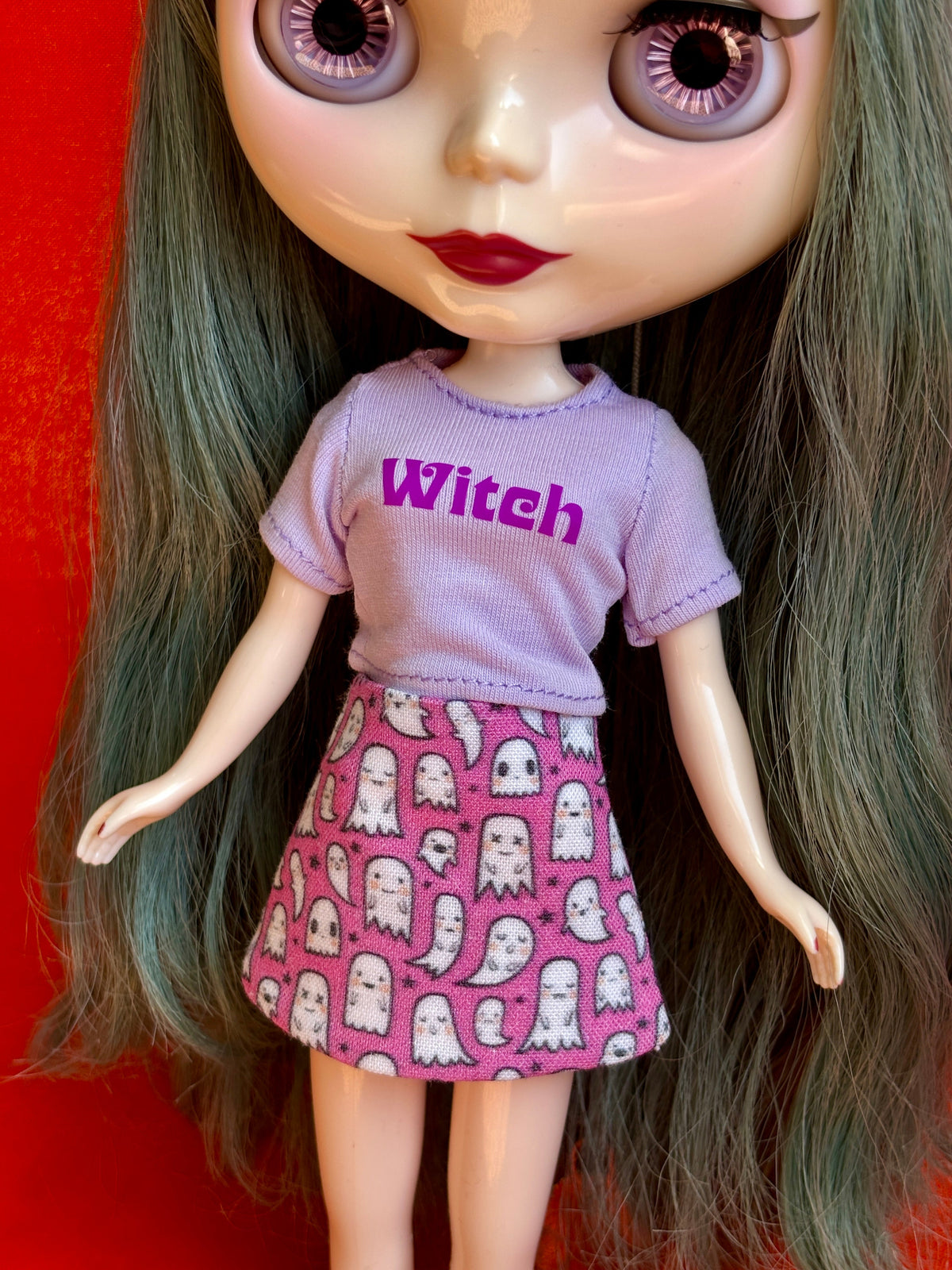 A close-up of a doll with large eyes and long dark green hair, wearing a light purple short-sleeve shirt with the word 'Witch' in darker purple text and a pink skirt covered in white ghost patterns, set against a solid red background.