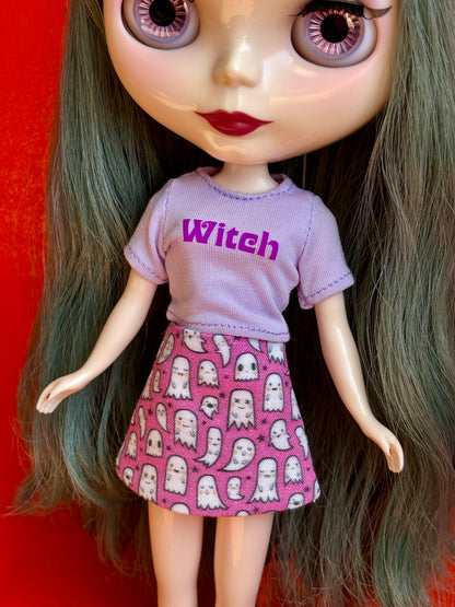 A close-up of a doll with large eyes and long dark green hair, wearing a light purple short-sleeve shirt with the word 'Witch' in darker purple text and a pink skirt covered in white ghost patterns, set against a solid red background.