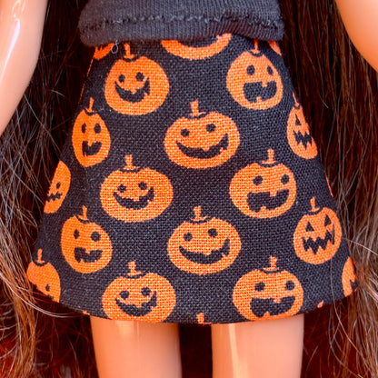 A close-up of a doll wearing a black skirt with an orange jack-o'-lantern pumpkin print, shown against the doll's dark hair and skin.