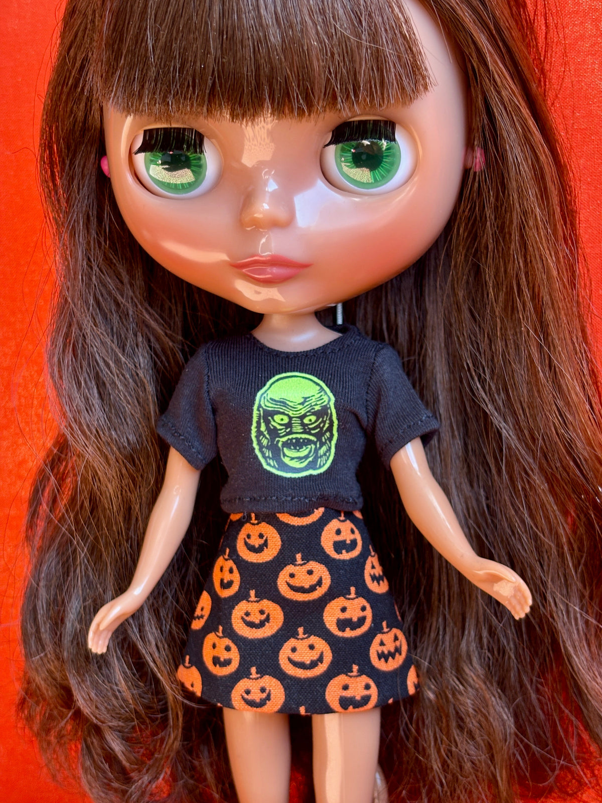 A close-up of a doll with long brown hair and large green eyes, wearing a black graphic T-shirt with a green monster face and a black skirt with an orange pumpkin print, set against a bright red background.