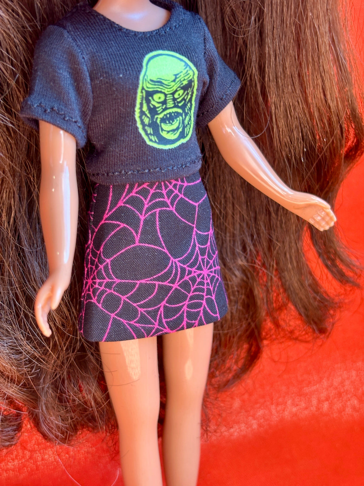 A doll with long brown hair wearing a dark gray short-sleeve top with a green monster face graphic and a dark skirt with a pink spiderweb pattern, posed against a bright orange-red background.