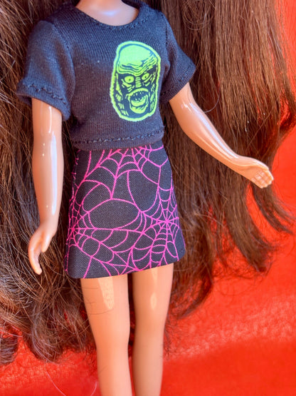 A doll with long brown hair wearing a dark gray short-sleeve top with a green monster face graphic and a dark skirt with a pink spiderweb pattern, posed against a bright orange-red background.