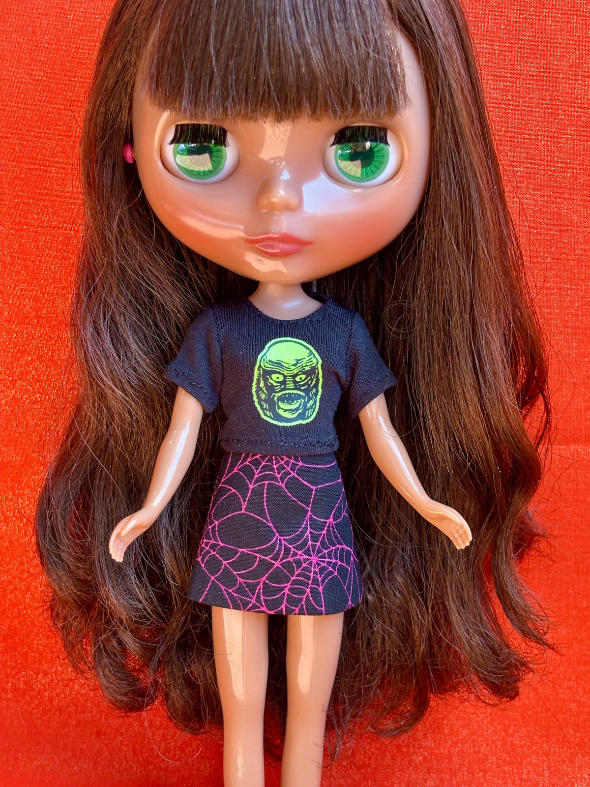 A doll with long brown hair and large green eyes, wearing a black t-shirt with a green monster face graphic and a black skirt with a pink spiderweb pattern, posed against a bright red background.