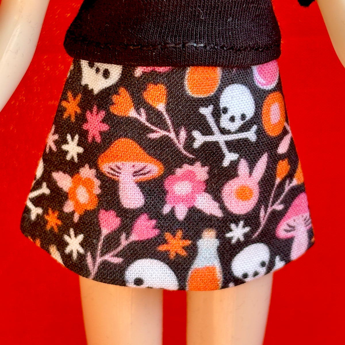 A close-up of a doll's lower body wearing a black skirt with a colorful Halloween pattern of orange mushrooms, pink flowers, white skulls, and orange pills, set against a solid red background.