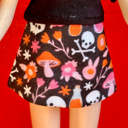 A close-up of a doll's lower body wearing a black skirt with a colorful Halloween pattern of orange mushrooms, pink flowers, white skulls, and orange pills, set against a solid red background.