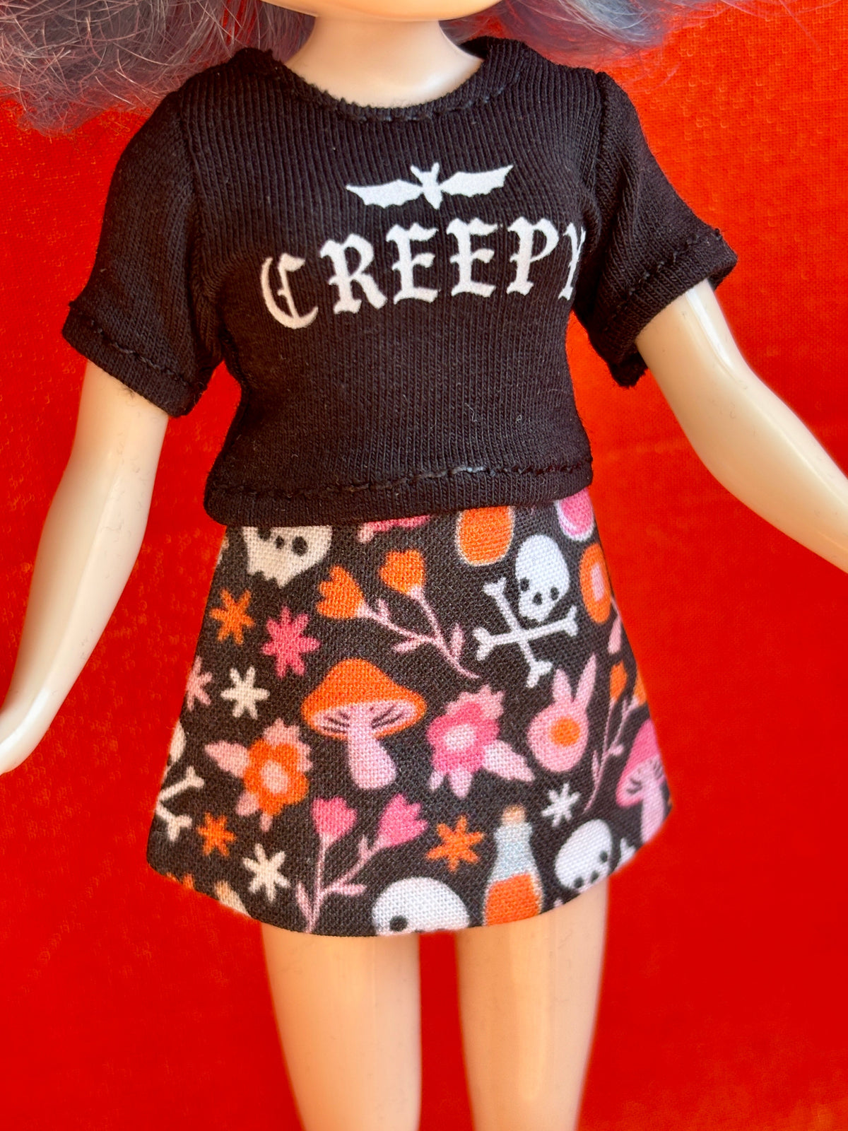 A doll or mannequin wearing a black short-sleeve top with the word 'CREEPY' in white gothic lettering and a black skirt with a colorful Halloween print of skulls, mushrooms, flowers, and bottles, set against a bright red background.