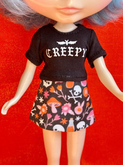 A doll or toy figure wearing a black short-sleeve top with the word 'CREEPY' in white gothic lettering, paired with a black skirt covered in a colorful Halloween-style print of skulls, mushrooms, flowers, and bats, posed against a bright red background.