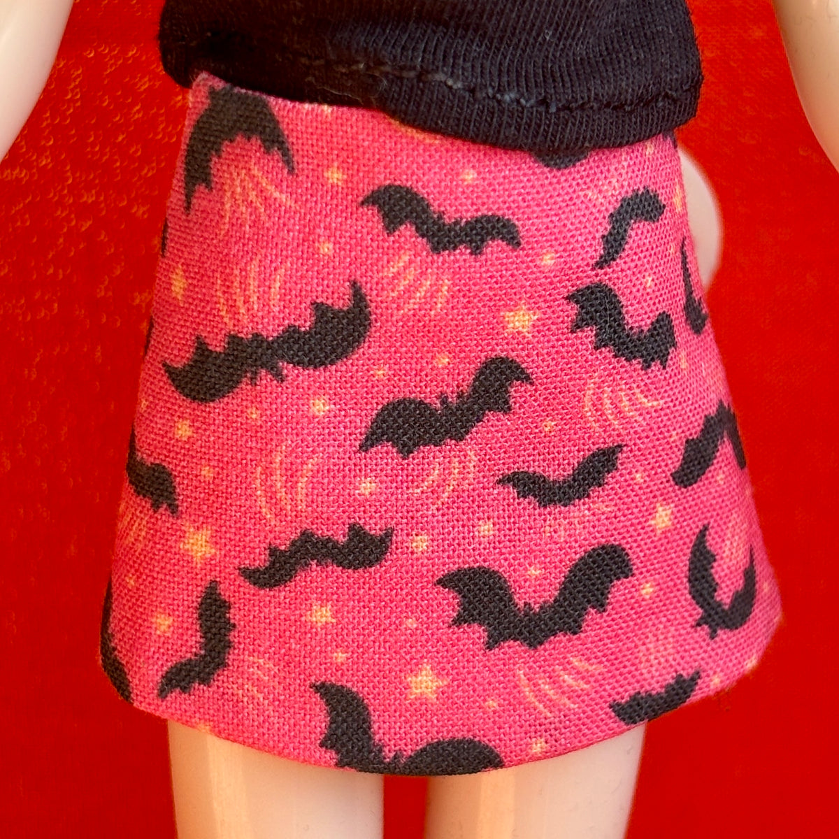 A close-up of a doll wearing a pink skirt with a black bat and moon pattern, paired with a black top, set against a bright red background.