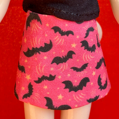 A close-up of a doll wearing a pink skirt with a black bat and moon pattern, paired with a black top, set against a bright red background.