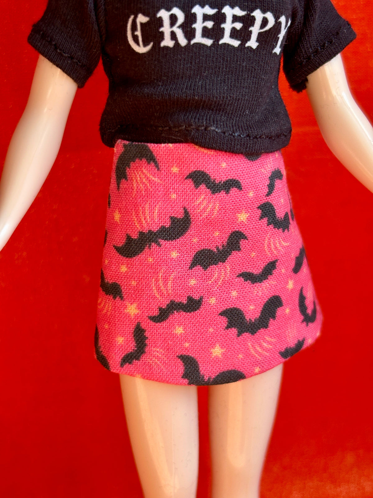 A doll or mannequin torso wearing a black short-sleeve top with the word 'CREEPY' in white gothic letters and a pink skirt with a black bat and yellow star pattern, set against a solid red background.