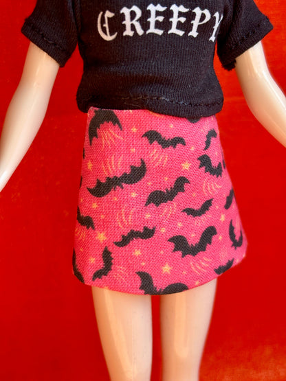 A doll or mannequin torso wearing a black short-sleeve top with the word 'CREEPY' in white gothic letters and a pink skirt with a black bat and yellow star pattern, set against a solid red background.