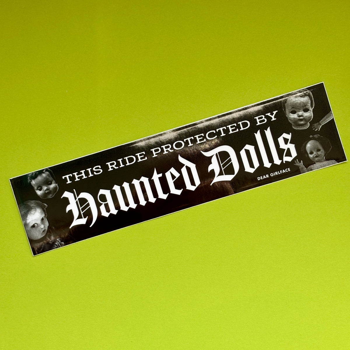 A rectangular bumper sticker with a black background and white gothic text reading 'This Ride Protected by Haunted Dolls,' accented with black-and-white photos of creepy dolls on the sides, shown on a bright lime green background.