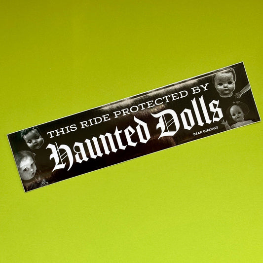 A rectangular bumper sticker with a black background and white gothic text reading 'This Ride Protected by Haunted Dolls,' accented with black-and-white photos of creepy dolls on the sides, shown on a bright lime green background.
