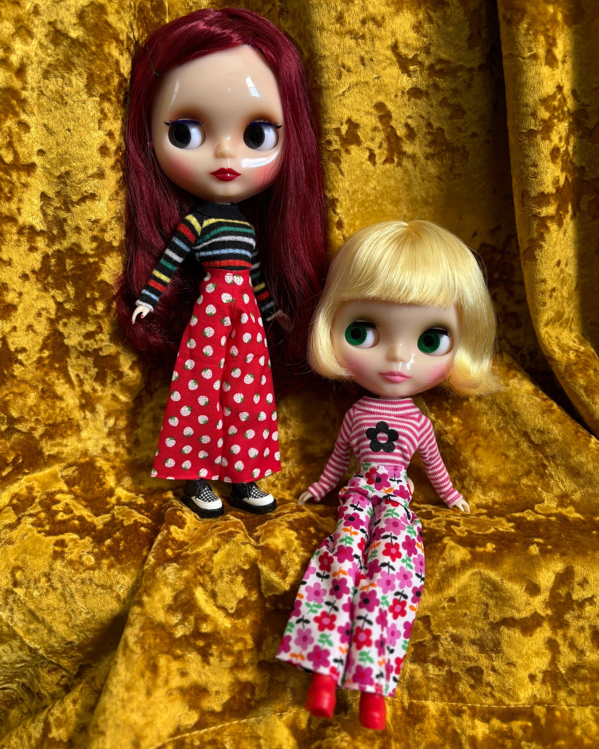 Two dolls with red hair and blonde hair, wearing colorful outfits, on a gold textured background.